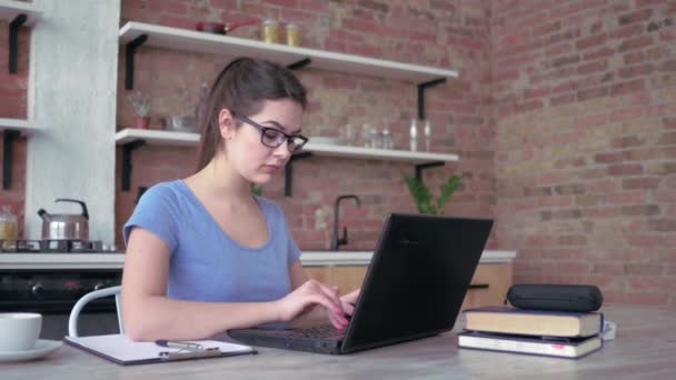 freelancer chica en gafas de escribir en el teclado del ordenador portátil y escribe notas en el portapapeles durante el trabajo en línea sentado en la mesa en casa
 - Imágenes, Vídeo