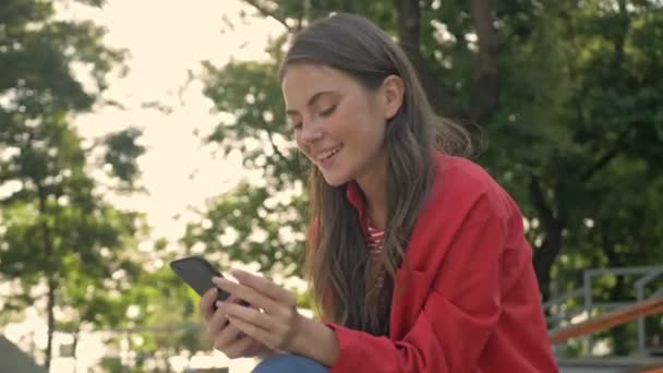 Cheerful cute young girl in red jacket smiling and using smartphone while sitting in the park - Video
