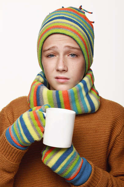 The sick boy holds a cup of tea, medicine, got sick. In a sweater, a cap, gloves, a disease, cold, flu, winter, fall. On a white background in studio. - Photo, Image