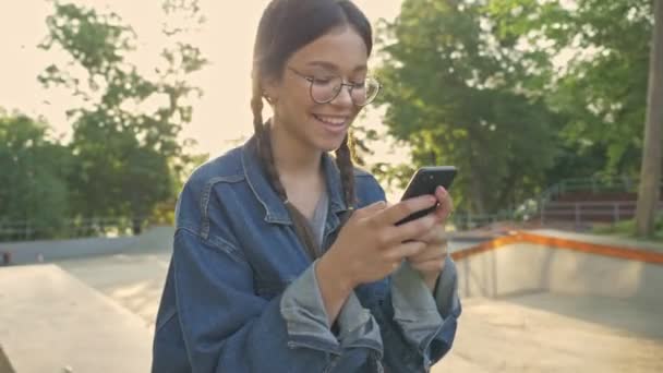 Attractive cheerful young brunette girl in denim jacket smiling and chatting on smartphone while sitting on her skateboard in skatepark - Imágenes, Vídeo