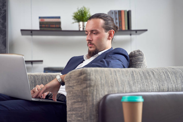 Side photo of brunet man with laptop sitting on sofa in room - Zdjęcie, obraz