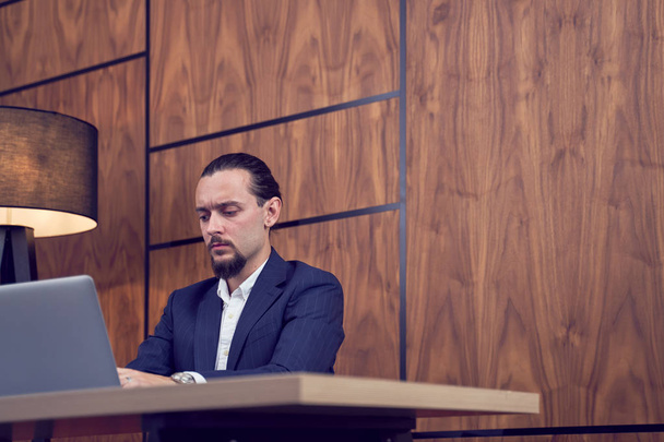 Photo of young businessman sitting at table with laptop on background of wooden wall. - Zdjęcie, obraz