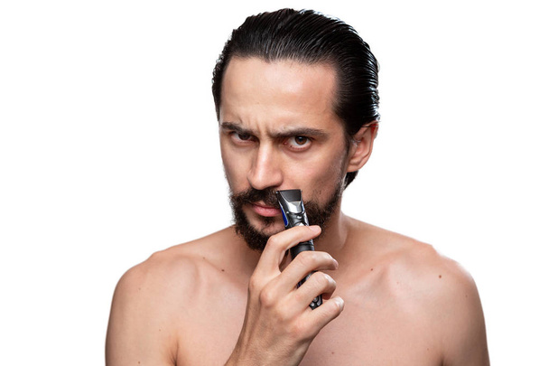 Красивый человек с усами использует электробритву, чтобы обрезать бороду стоя голые изолированы на белом фоне. Концепция утреннего лечения и бритья. Пора подстричь бороду. Утренняя программа
 - Фото, изображение