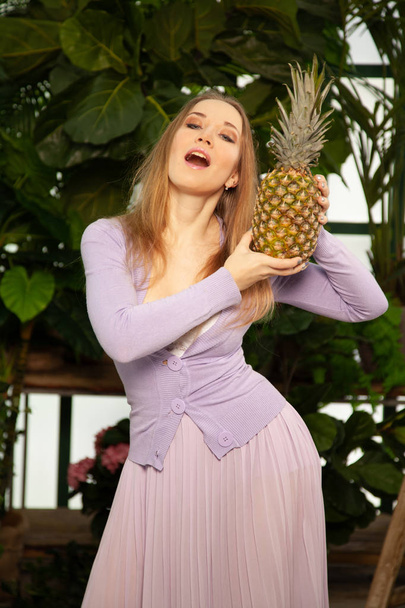 beautiful girl in a purple dress with a pineapple in her hands in the greenhouse poses among the plants and enjoys the clean air and harmony from communicating with nature - Photo, Image