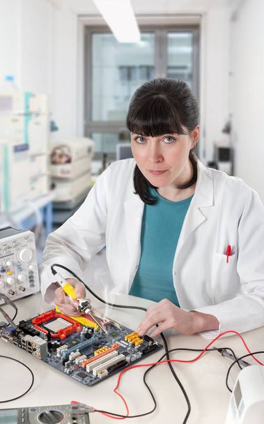 Young female tech soldering a circuit board looks at the viewer - Фото, зображення