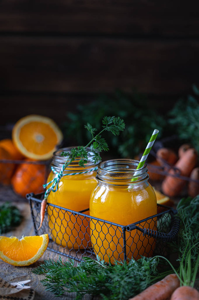 Egészséges étel. Sárgarépa és sárgarépalé narancssárgával üvegedényben, fémkosárban, sötét fa alapon - Fotó, kép