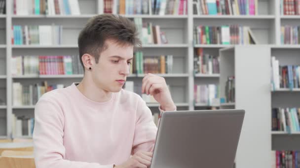 Młody człowiek wygląda troskliwie podczas pracy nad laptopem w bibliotece - Materiał filmowy, wideo