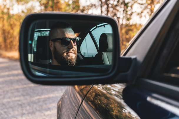 Cute, bearded guy driving a car, reflection in car rearview mirror. - Фото, изображение