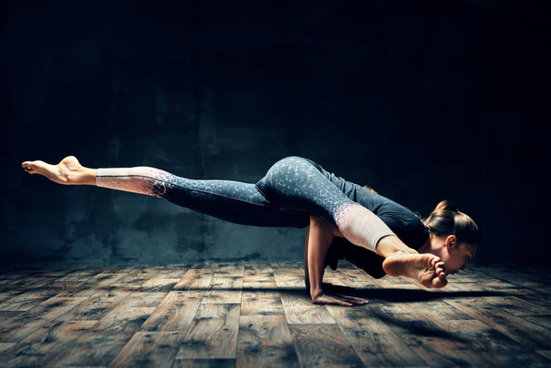 Young woman practicing yoga doing hurdler pose in dark room. Eka Pada Koundinyasana.  - Fotografie, Obrázek