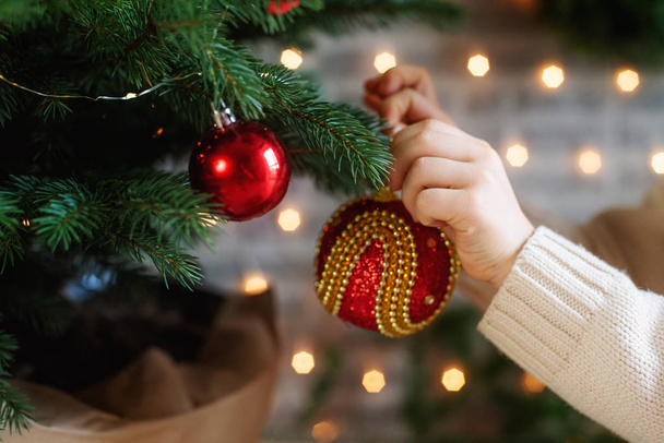Ένα παιδί διακοσμεί ένα χριστουγεννιάτικο δέντρο. Χριστουγεννιάτικη μπάλα στο χέρι ενός παιδιού. - Φωτογραφία, εικόνα