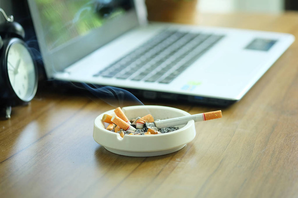 Muchos cigarrillos en cenicero blanco en el escritorio con computadora portátil. Fumar para aliviar el estrés en el trabajo pero poco saludable
 - Foto, imagen