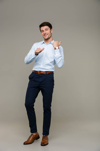 Image of joyful handsome man wearing formal clothes smiling and pointing fingers backward at copyspace - Фото, изображение
