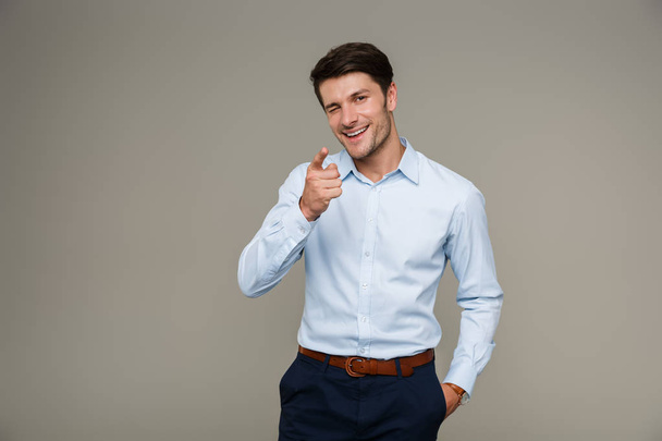 Image of cheerful brunette man wearing formal clothes smiling and pointing finger at camera while winking - Foto, afbeelding