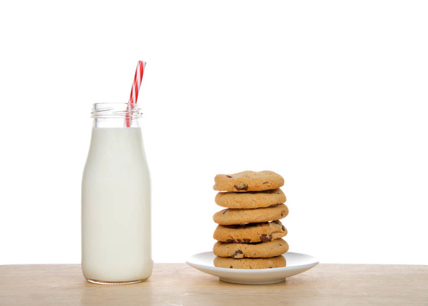 Small glass bottle of fresh milk with candy cane pattern striped straw on table next to stack of homemade chocolate chip cookies on a plate.  - Фото, изображение