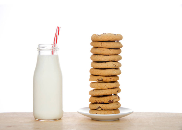 Kleine glazen fles verse melk met suikerriet patroon gestreept stro op tafel naast stapel zelfgemaakte chocolade chip koekjes op een bord. Een dozijn koekjes onzeker gestapeld. - Foto, afbeelding