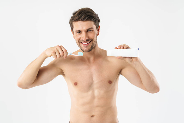 Cheery positive young naked man brushing his teeth isolated over white background. - Fotografie, Obrázek