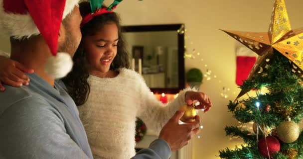 Side view close up of a mixed race father with his young daughter in their sitting room at christmas, the man is wearing a santa hat and holding his daughter while she decorates the christmas tree - 映像、動画