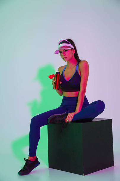 Serious young sports woman posing isolated with led flash lights in cap holding bottle with water. - Foto, Imagen