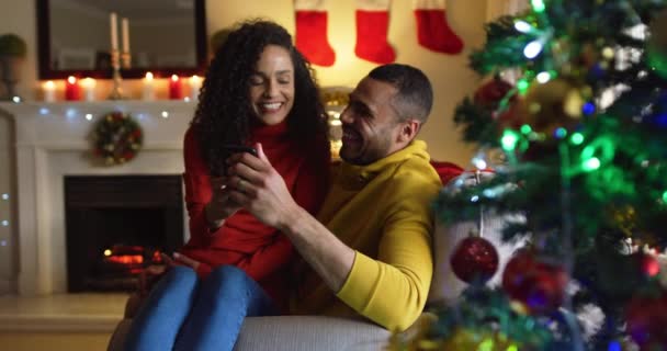 Front view of a mixed race couple sitting on a sofa in their sitting room at Christmas using a smartphone togther, the woman sitting on the lap of the man - 映像、動画