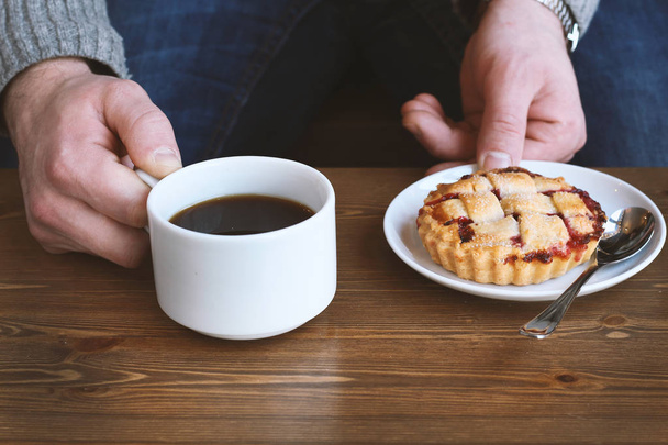 man in grey sweater drinks black lungo americano coffee in winter autumn in coffee shop eating classic american home-made cherry pie on white plate on wooden table. Fall mood instagram style - Fotó, kép