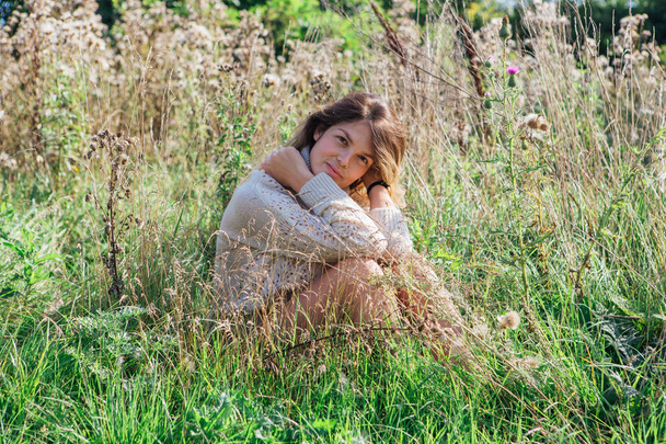 Happy young beautiful woman in sweater and underpants sitting with bare foot in a field of dry agrimony - Фото, зображення