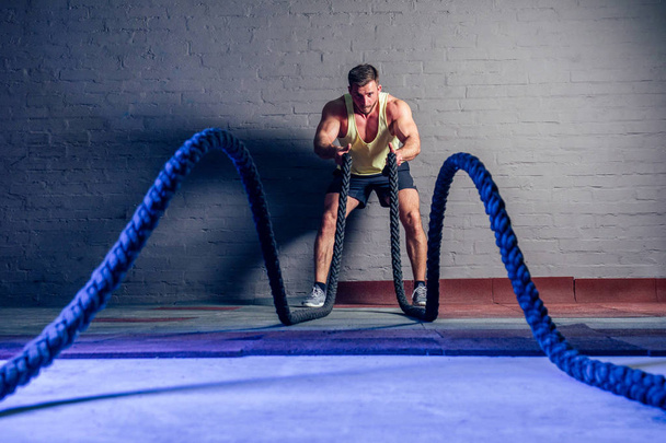 young and handsome male athlete man doing exercises for the muscles of the body with a battle ropes rope in fitness gym - Foto, immagini