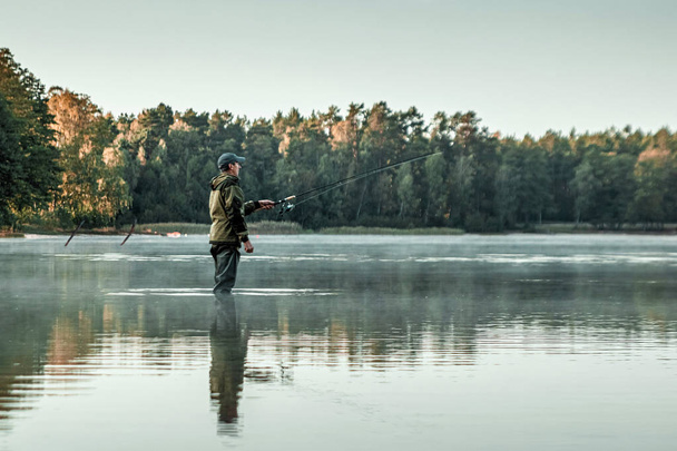 A male fisherman on the lake is standing in the water and fishing for a fishing rod. Fishing hobby vacation concept. Copy space. - Photo, image