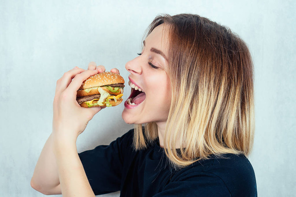 young and attractive blonde woman in black T-shirt and measuring tape eating a high-calorie burger. concept of harmful fast food and diet - Photo, Image