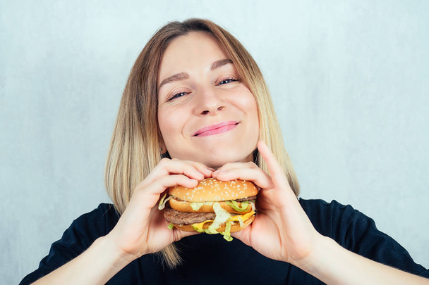 young and attractive blonde woman in black T-shirt and measuring tape enjoying a high-calorie burger. slimming and diet concept - Foto, afbeelding