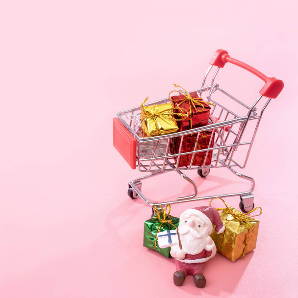 Christmas shopping concept, mini red shop cart trolley with Santa Claus toy and gift box isolated on pale pink background, blank copy space, close up - Foto, imagen