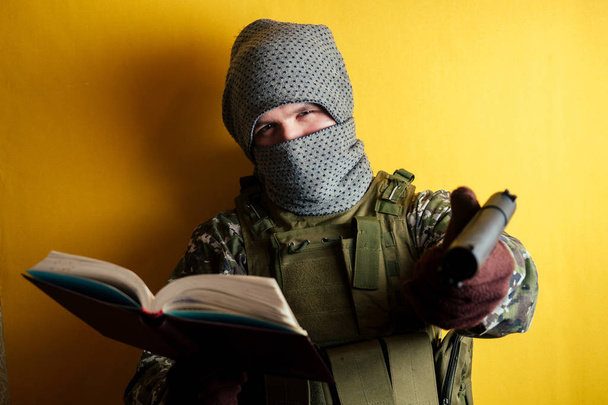 a terrorist man in a camouflage and a mask is holding a red book and weapons on a yellow background. the concept of anonymity and terrorism. - Foto, Imagem