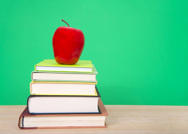 Hard bound books with various colored covers stacked on light wood table with red apple on top, dark green background similar to old fashioned chalk board. Back to school concept. - 写真・画像