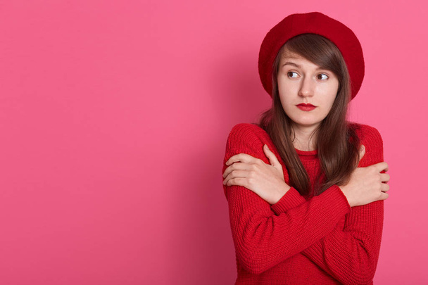 Half length shot of brunette woman trembles from cold, has fever, has crossed hands over chest, wears red clothes and beret, isolated over rosy background. Copy space for advertisment or promotion. - Photo, image