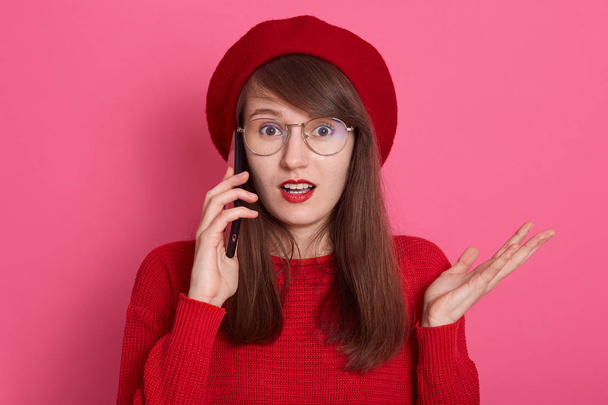 Photo of astonished girl wearing red beret, sweater,spectacles, talking on smartphone with her friend, spreading hand aside, keeps mouth opened, model posing isolated over rose background in studio. - Fotografie, Obrázek