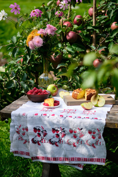 Egy szelet almás pitéből egy tányéron fekszik. Rusztikus csendélet: napos fényes nap, almás pite egy almafa alatt egy régi fa asztalon, gyümölcsök és bogyók, virágok. - Fotó, kép