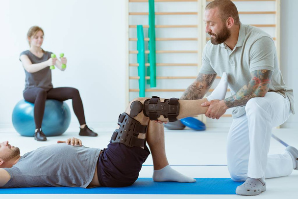 Teenage girl after car accident with orthosis on the leg exercises on blue mat in the company of physiotherapist - Foto, afbeelding