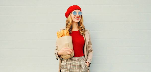 Portrait happy young smiling woman looking away holding grocery shopping paper bag with long white bread baguette wearing red beret on blank gray wall background - Φωτογραφία, εικόνα