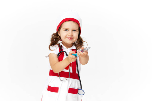 Happy cute little child girl in doctor costume and glasses holding stethoscope on white background. Holding a toy syringe in her hands. Vaccination concept - Photo, image