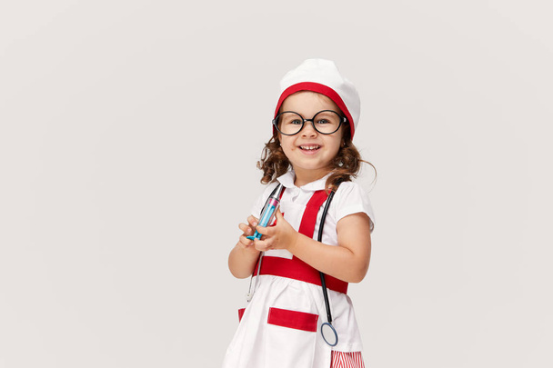 Joyeux petit enfant fille mignonne en costume de médecin et lunettes tenant stéthoscope sur fond blanc. Tenir une seringue dans ses mains. Concept de vaccination
 - Photo, image