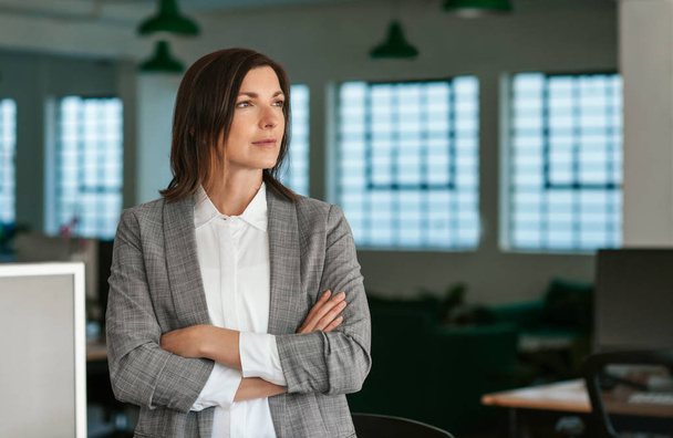 Focused businesswoman looking deep in thought while standing with her arms crossed in a large modern office - Фото, зображення