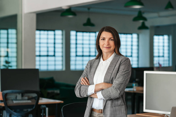 Portrait of a smiling businesswoman standing with her arms crossed by her desk in a large modern office in the late afternoon - Foto, Bild