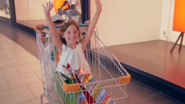 season of discounts, laughing children are having fun in shopping trolleys in mall and go past shop windows of boutiques - Footage, Video