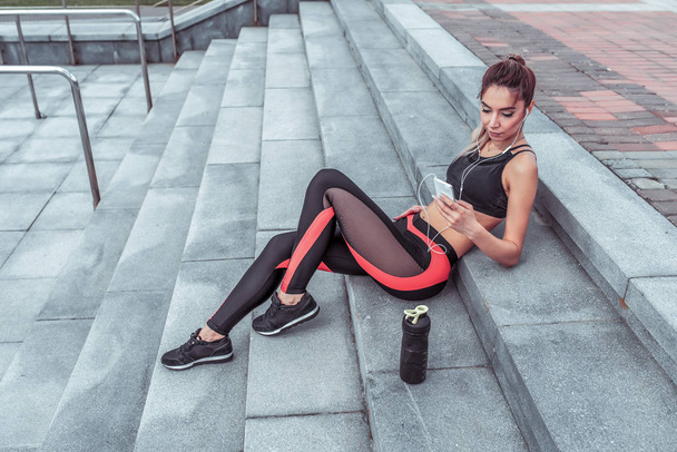 Beautiful girl, summer autumn and spring city, resting active training, holding telephone listening music headphones, watching videos reading message active fitness lifestyle, outdoor sports exercise - Photo, Image