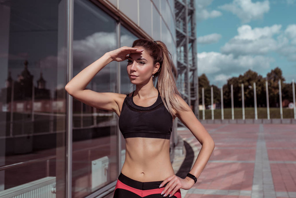 Beautiful girl athlete, covers distance into distance with hand from sun, glass windows background, summer spring city, free space motivation, sportswear, active fitness lifestyle, outdoor exercise. - Foto, imagen