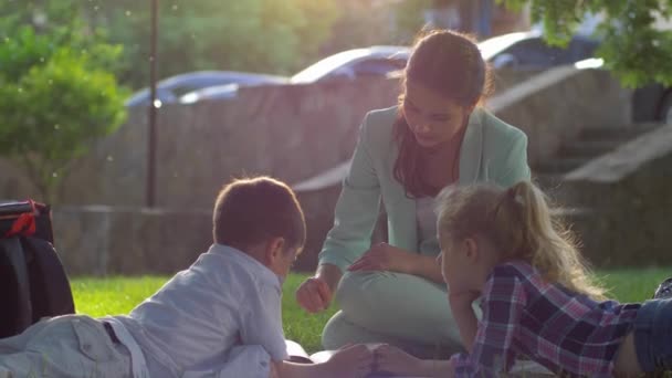 educatieve lessen, dame leraar lees boek voor kleine jongen en meisje zitten op groen gras in de natuur in zonnig licht - Video