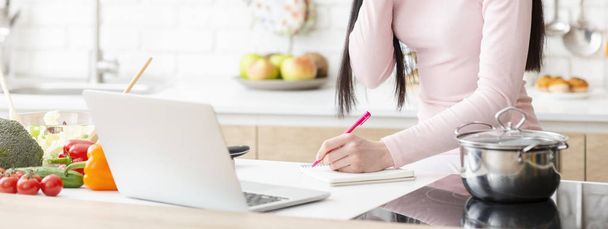 Giovane donna che guarda webinar sul computer portatile e prendere appunti in cucina
 - Foto, immagini