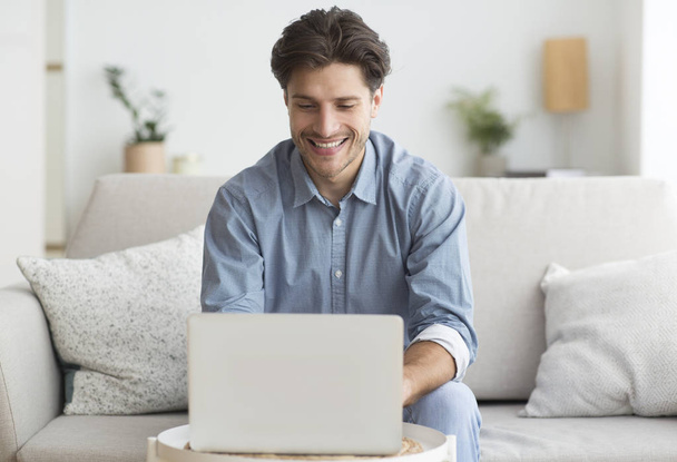 Улыбающийся человек работает на ноутбуке, сидя на диване дома
 - Фото, изображение
