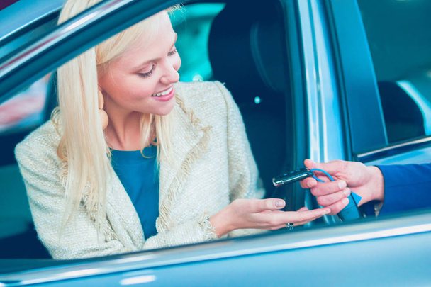 Beautiful young woman is getting key and smiling while sitting in a new car in dealership - 写真・画像