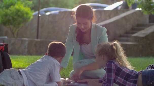 lezen boeken buiten, vrolijke vrouwelijke leraar lees boek voor kleine jongen en meisje zitten op groen gras op open lucht in zonnig licht - Video