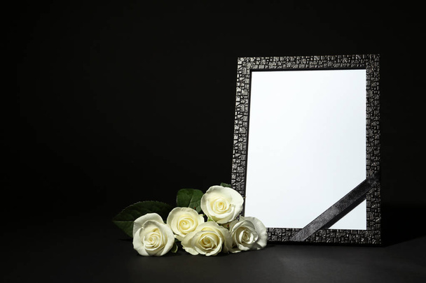Funeral photo frame with ribbon and white roses on dark table against black background. Space for design - Photo, image